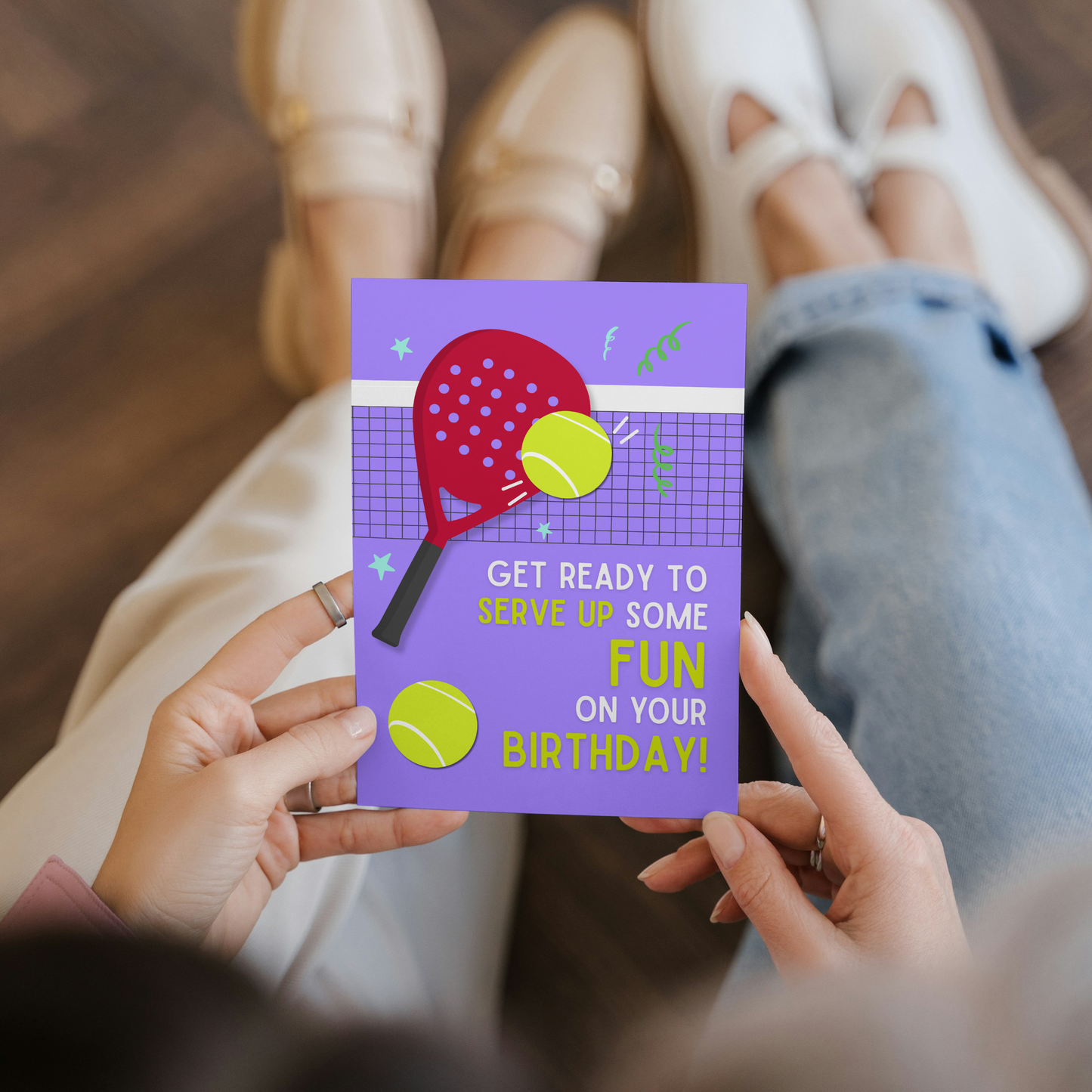Person holding a birthday card with a tennis theme on a wooden floor.